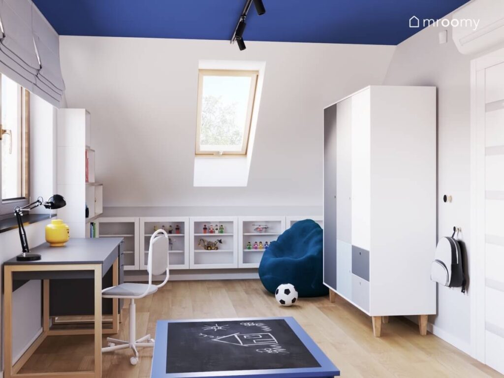 Poddaszowy pokój dla chłopca z ciemnoniebieskim sufitem dużą szafą z frontami w różnych kolorach ciemnoniebieską pufą i szafkami pod skosem