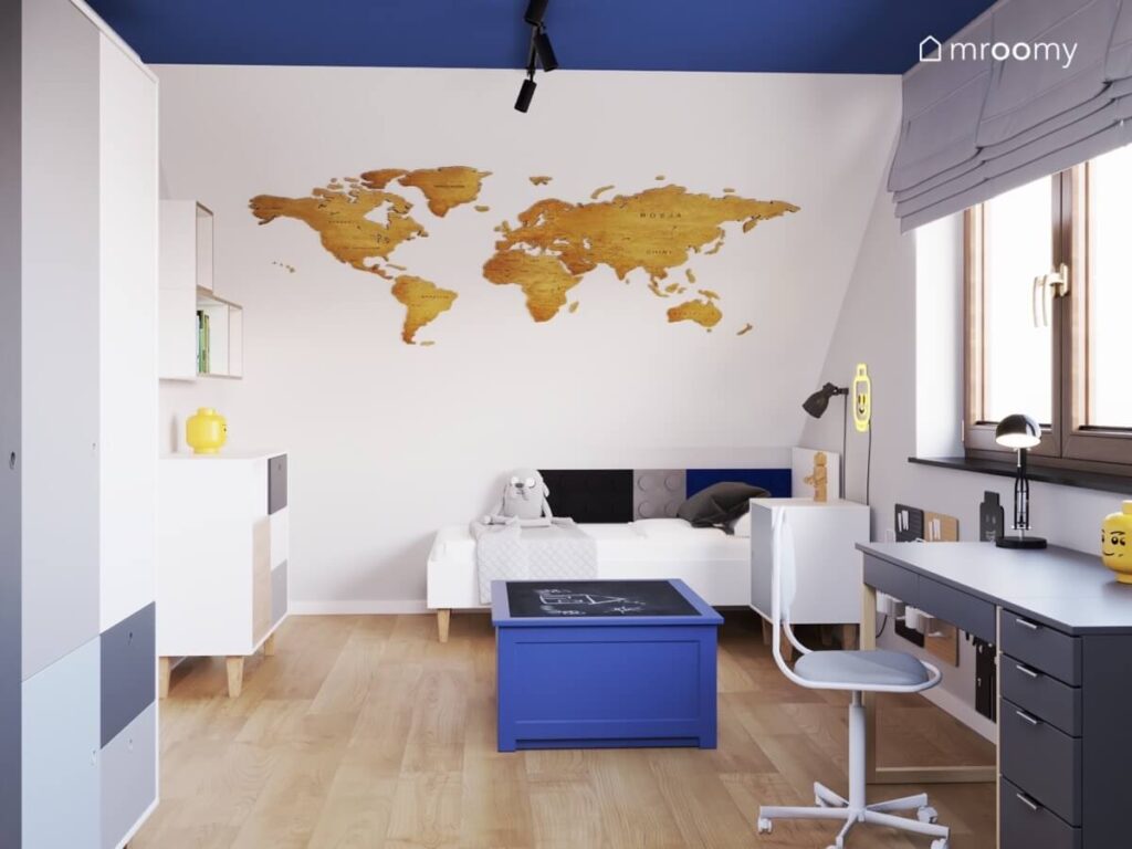 Biały skos w poddaszowym pokoju chłopca a na nim drewniana mapa świata a pod spodem proste białe łóżko uzupełnione panelami ściennymi w kształcie klocków