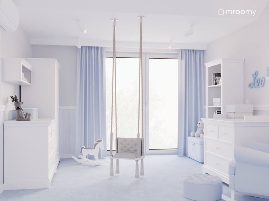Białe meble niebieskofioletowe zasłony konik na biegunkach oraz wisząca huśtawka z frędzlami w pokoju małego chłopca