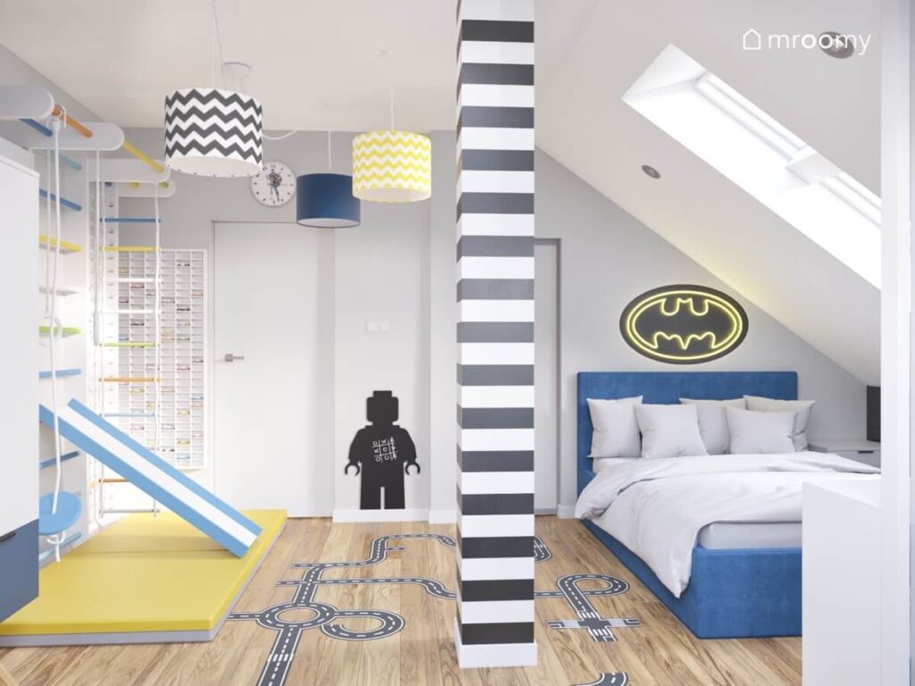 Niebieskie tapicerowane łóżko a nad nim ledon w kształcie symbolu Batmana czarnobiały filar a na suficie lampa z trzema różnymi filarami w poddaszowym pokoju chłopca
