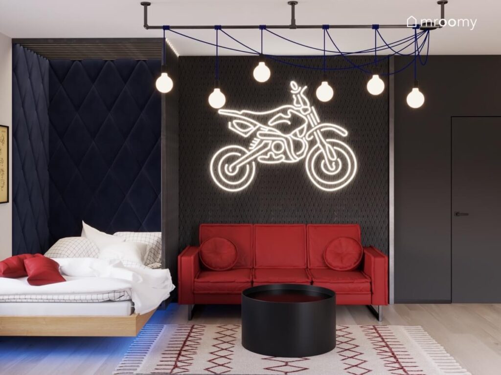 Wyłożona miękkimi panelami strefa spania w pokoju nastolatka a obok czerwona sofa ze stolikiem a nad nią ledon motocykl i luźno wiszące żarówki