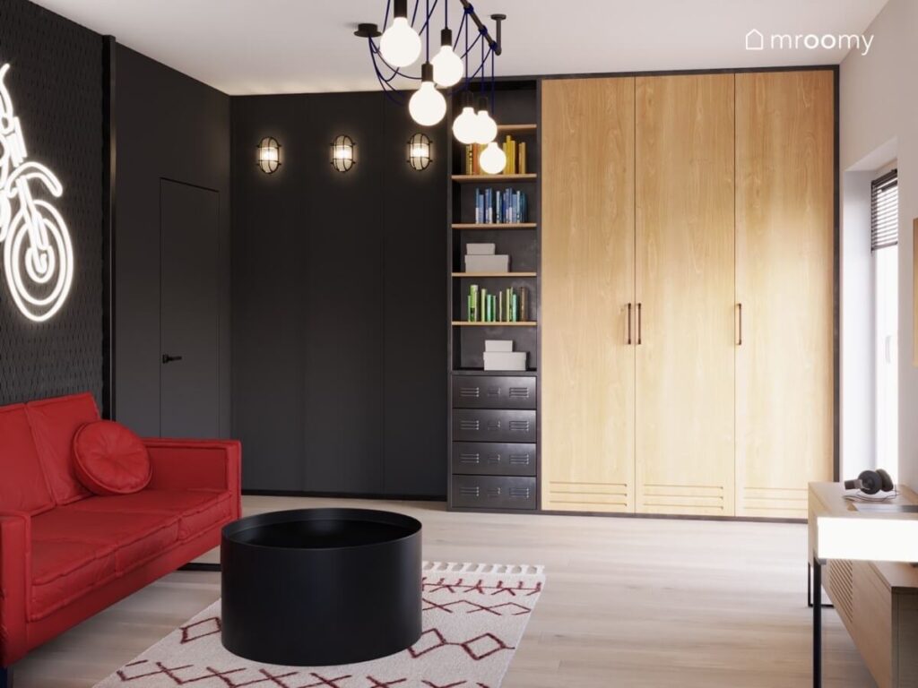Wysoki czarny regał oraz drewniana szafa a na środku czerwona sofa i czarny stolik w pokoju dla nastolatka