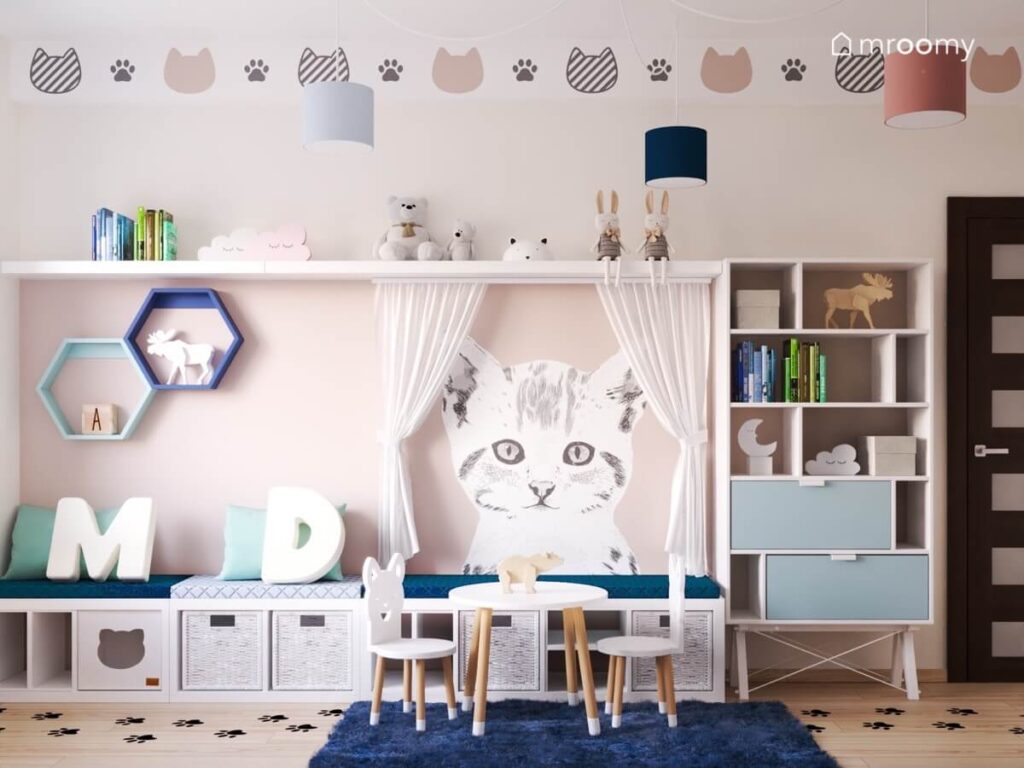 Beżowa ściana w pokoju dla rodzeństwa a na niej duży regał z niebieskimi szufladami długie siedzisko półki oraz naklejka z kotem