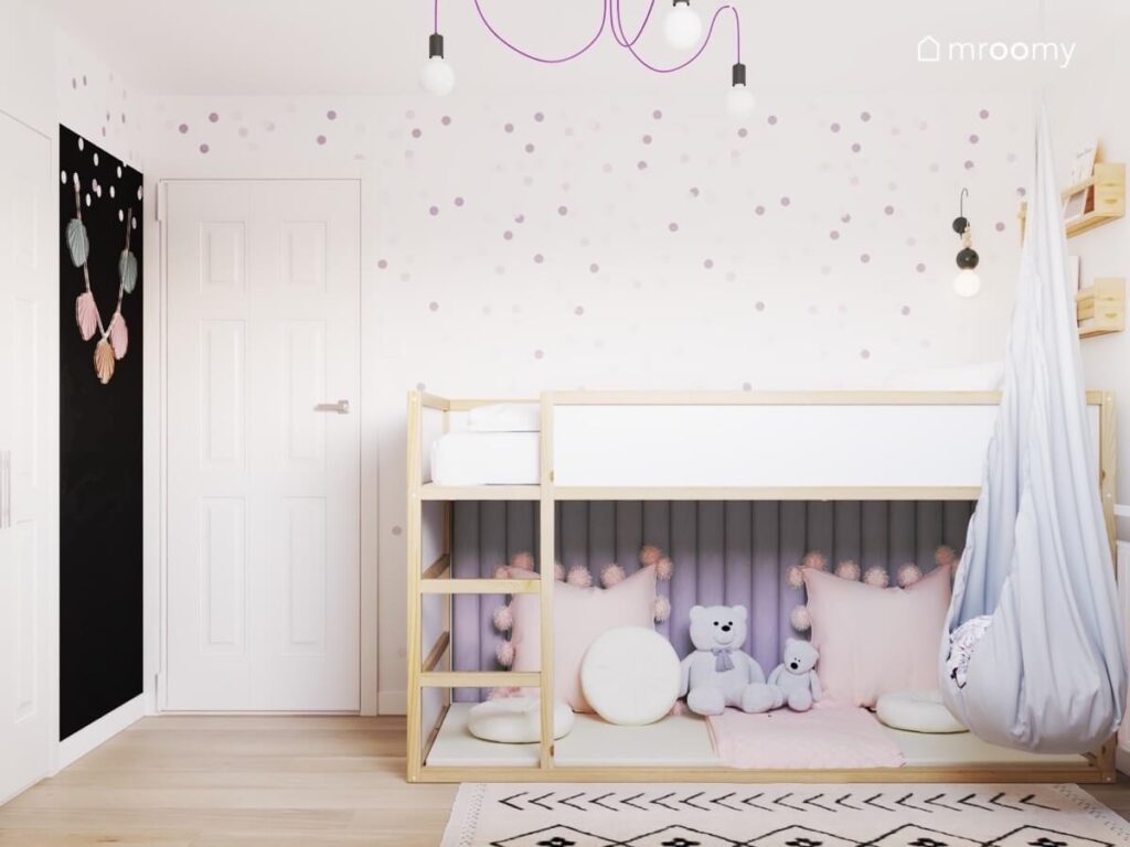 Biało drewniane łóżko na antresoli z przytulnym kącikiem pod spodem na ścianie kropki a u sufitu lampa na fioletowym zawieszeniu w pokoju dziewczynki w wieku szkolnym