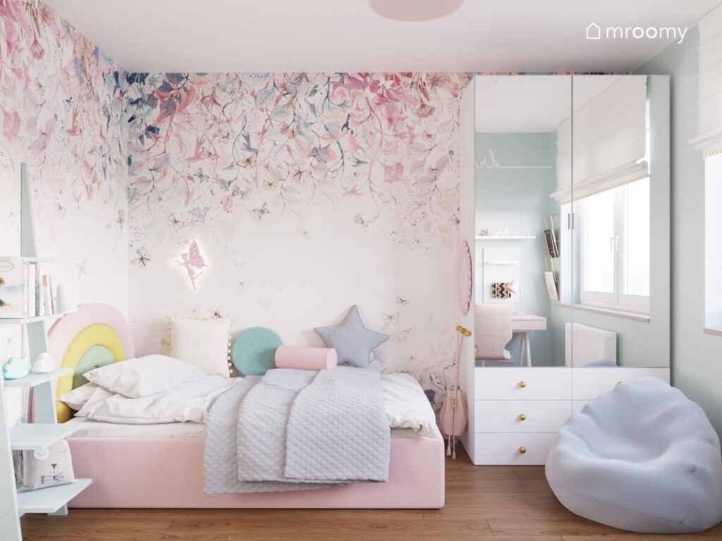 Różowe tapicerowane łóżko z zagłówkiem w formie tęczy obok biała szafa z lustrzanymi frontami oraz jasna pufa a na ścianie roślinna tapeta w pokoju dziewczynki