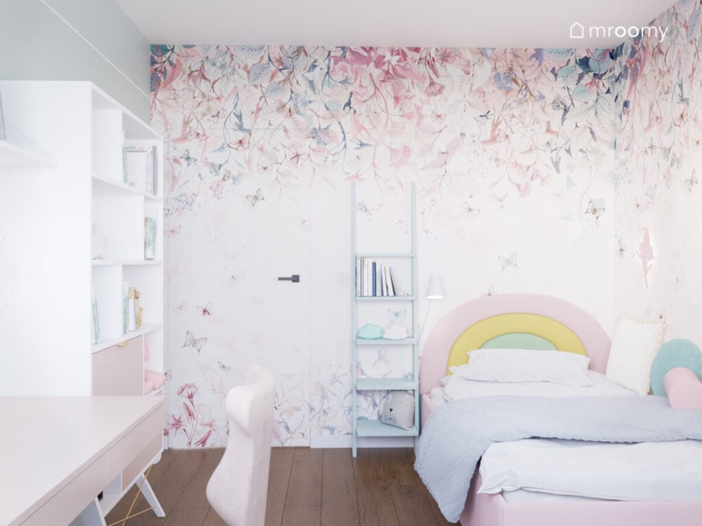 Jasny pokój dla dziewczynki z tapicerowanym łóżkiem z zagłówkiem w formie tęczy regałem drabinką oraz tapetą w kwiaty