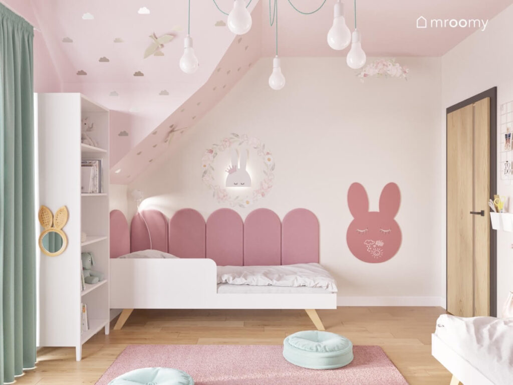 Strefa spania w poddaszowym pokoju dziewczynek a w niej białe łóżko uzupełnione różowymi panelami lampka oraz tablica kredowa królik a na skosie naklejki