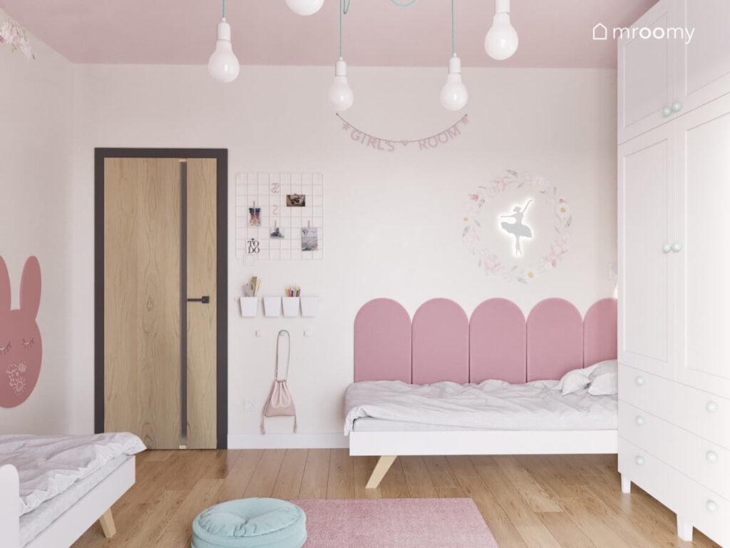 Proste łóżko uzupełnione różowymi panelami ściennymi a nad nim lampka baletnica a obok organizery ścienne w pokoju dla dwóch dziewczynek