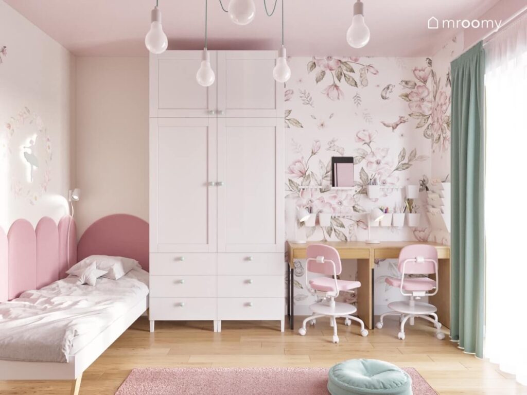 Biało różowy pokój dla dwóch dziewczynek z prostym łóżkiem uzupełnionym miękkimi panelami dużą szafą oraz kwiecistą strefą nauki z drewnianymi biurkami