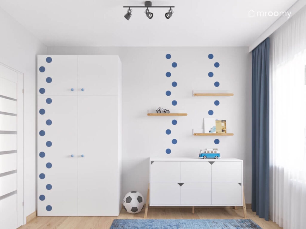 Duża biała szafa z niebieskimi kropkami obok biała komoda a na ścianie drewniane półeczki i również niebieskie kropki w pokoju chłopca