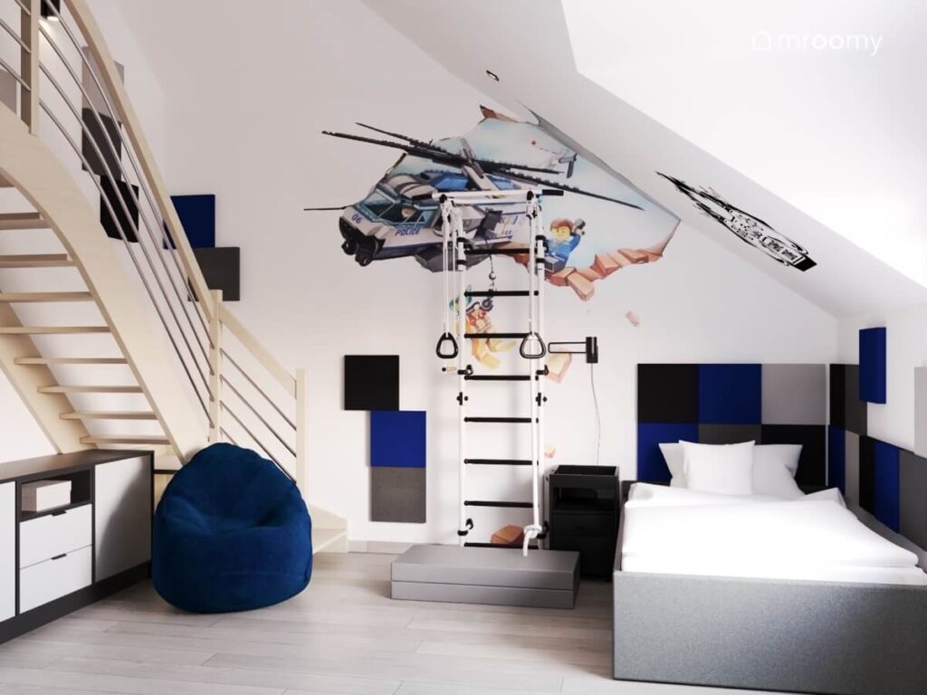 Biały poddaszowy pokój dla chłopca z szarym łóżkiem uzupełnionym miękkimi panelami drabinką gimnastyczną granatową pufą oraz naklejką z helikopterem