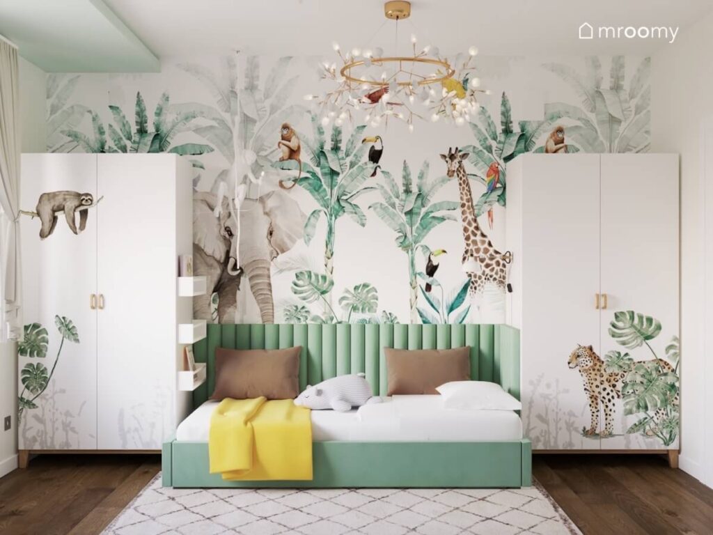 Ściana pokryta tapetą w dzikie zwierzęta a na niej dwie białe szafy ze zwierzęcymi naklejkami oraz zielone tapicerowane łóżko w pokoju dziewczynki