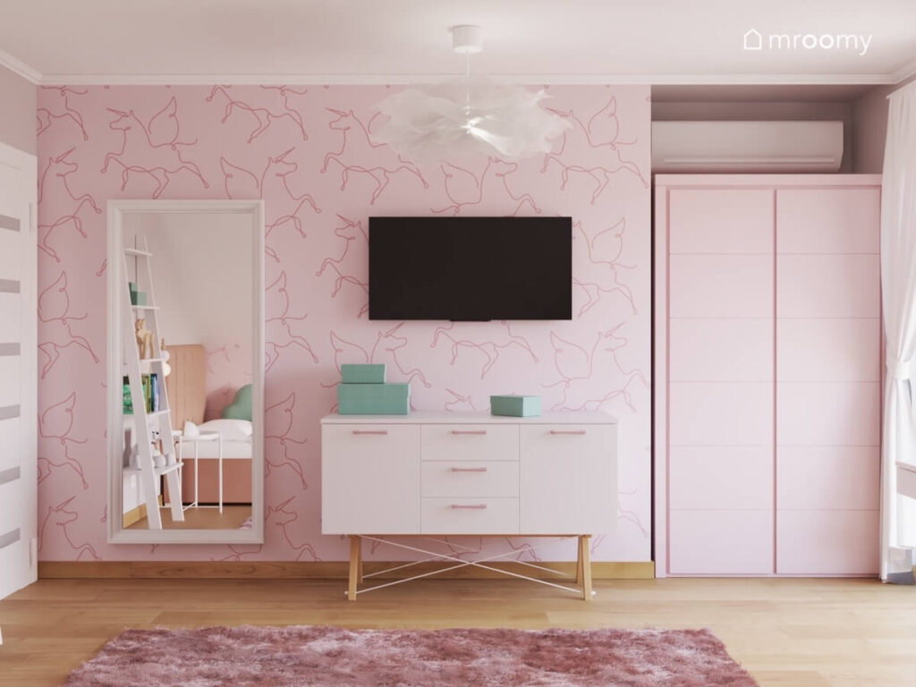 Ściana w pokoju dziewczynki pokryta tapetą w latające jednorożce a na niej duże prostokątne lustro telewizor i szafka na drewnianych nogach