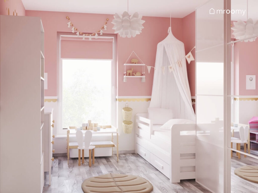 Różowo biały pokój dla dziewczynki a w nim białe meble łóżko uzupełnione baldachimem oraz ozdobna lampa na suficie