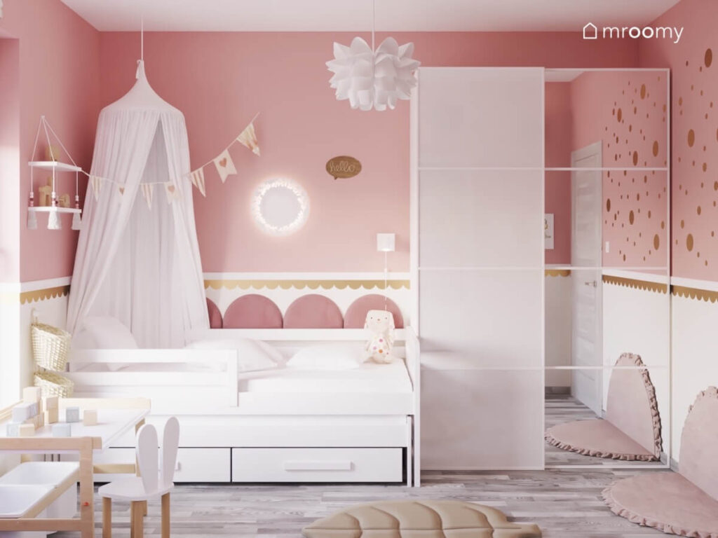 Strefa spania w pokoju malutkiej dziewczynki a w niej łóżko z szufladami i baldachimem na ścianie panele ścienne i biała lampka a obok szafa z lustrzanym frontem