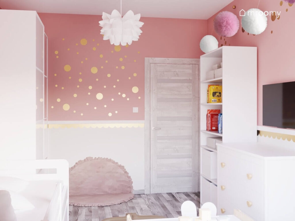 Biało różowe ściany w pokoju małej dziewczynki a na nich złote kropki oprócz tego biały regał pompony i ozdobna lampa