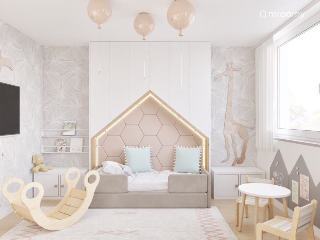 Tapicerowane łóżko w formie domku obok bujak na suficie lampy balony a na ścianie tapeta w liście i żyrafa ze sklejki w pokoju chłopca i dziewczynki
