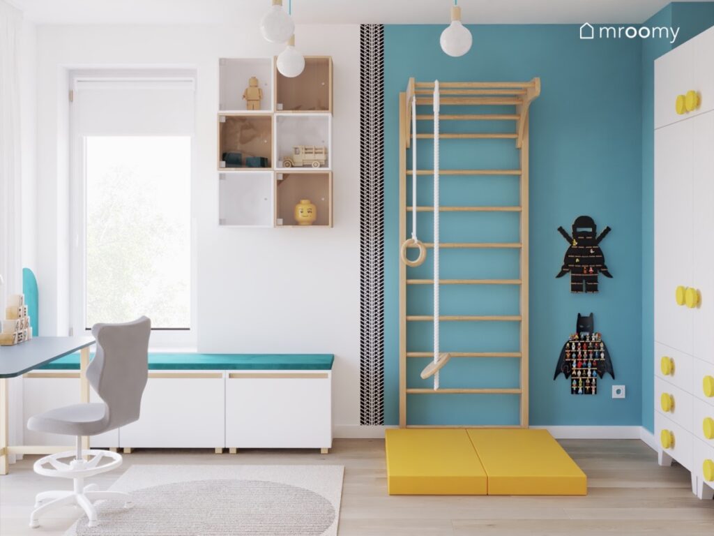 Biało niebieska ściana w pokoju chłopca na niej małe szafki drabinka gimnastyczna oraz półeczki na ludziki Lego