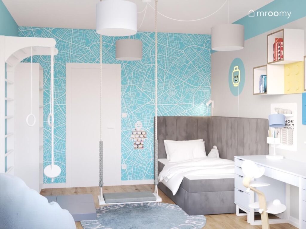 Błękitna tapeta z wzorem mapy szare tapicerowane łóżko oraz huśtawka wisząca w pokoju dla chłopca