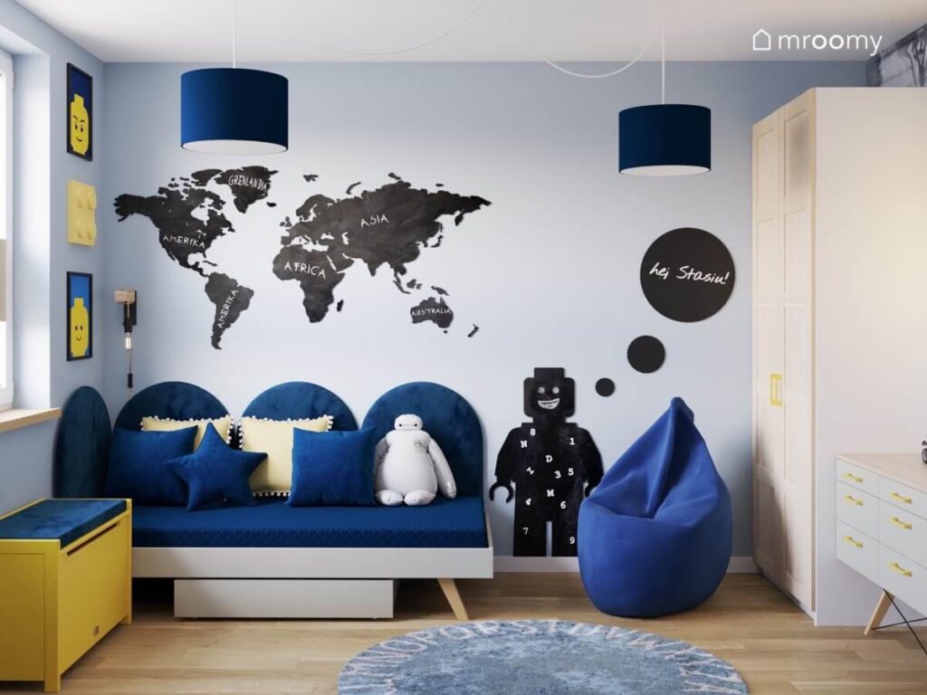 Strefa spania w pokoju dla chłopca a w niej proste łóżko uzupełnione miękkimi panelami nad nim tablica kredowa w kształcie mapy świata obok tablica ludzik i miękka pufa