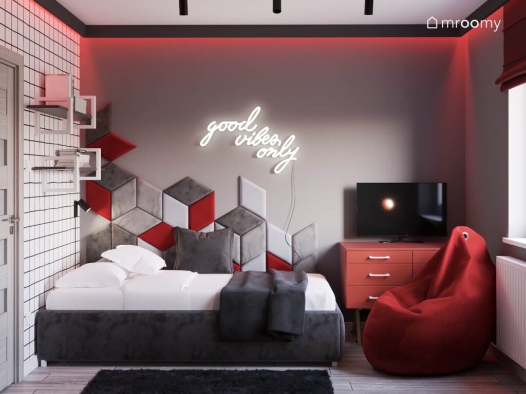 Strefa spania w szarym pokoju nastolatka a w niej szare tapicerowane łóżko kompozycja z paneli ściennych w różnych odcieniach oraz ledon a obok czerwona szafka i pufa