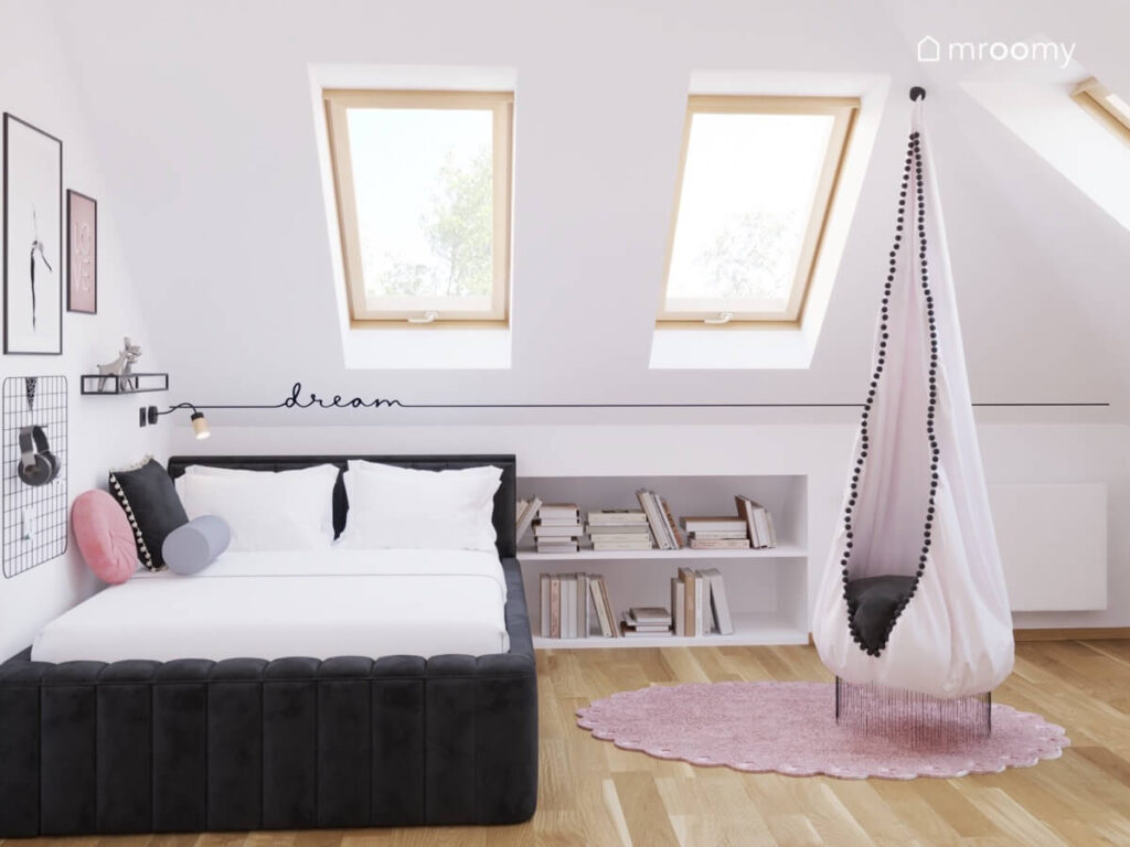 Czarne tapicerowane łóżko niski regał oraz fotel kokon na podłodze różowy dywan a na ścianie naklejka w poddaszowym pokoju dziewczynki