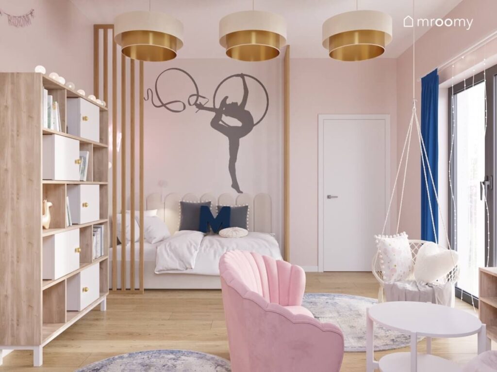 Biało drewniany regał łóżko uzupełnione panelami ściennymi różowa sofa i fotel wiszący na ścianie naklejka gimnastyczna a u sufitu biało złote lampy w pokoju dziewczynki