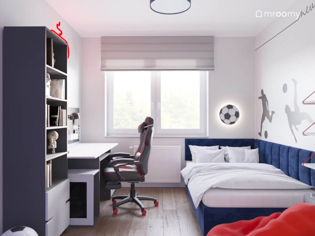 Jasny pokój dla chłopca z ciemnoniebieskim tapicerowanym łóżkiem biało szarym biurkiem i regałem oraz piłkarskimi dodatkami