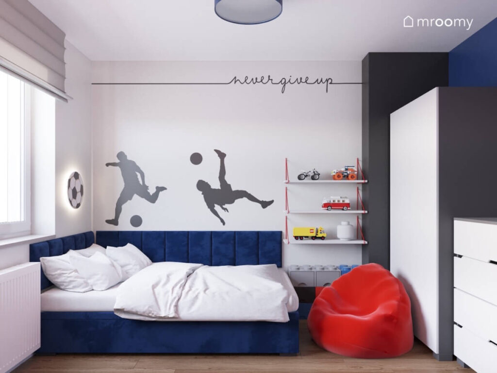 Strefa spania w pokoju chłopca z niebieskim tapicerowanym łóżkiem czerwoną pufą oraz naklejkami ściennymi w kształcie piłkarzy i napisu