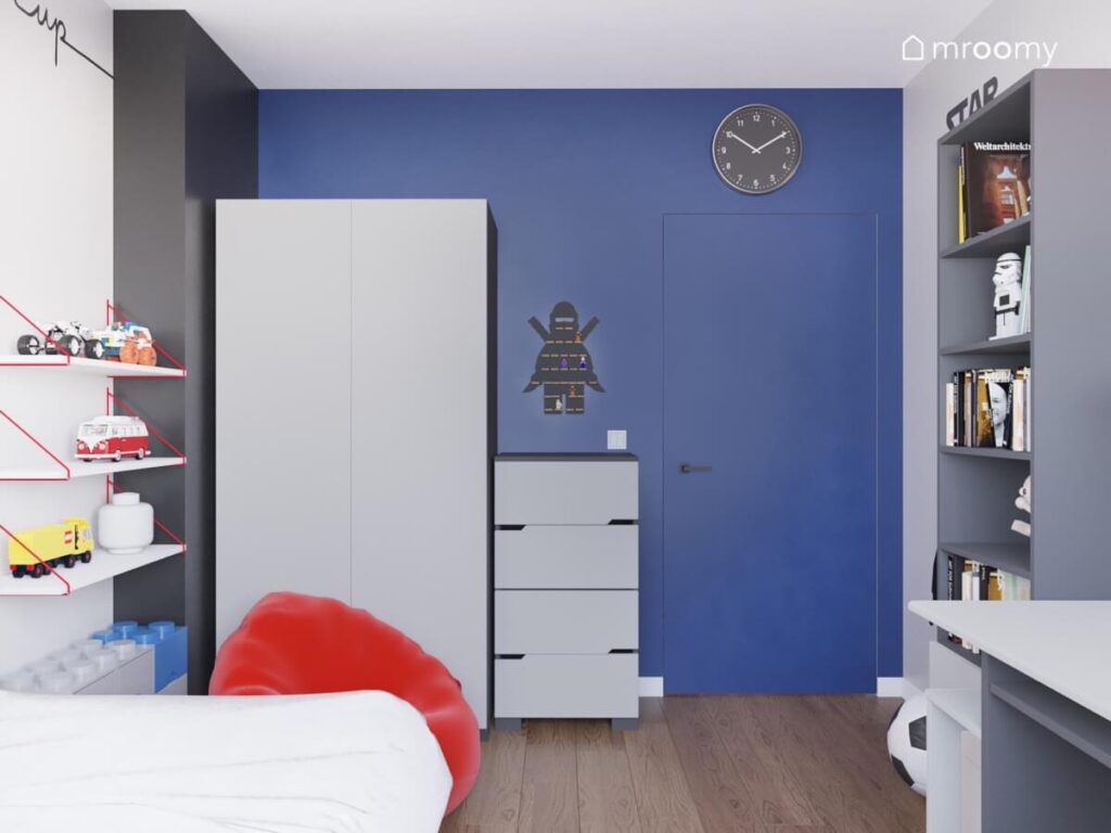 Niebieska ściana w pokoju chłopca a na niej szafa dwudrzwiowa komoda półka na ludziki Lego oraz czarny zegar