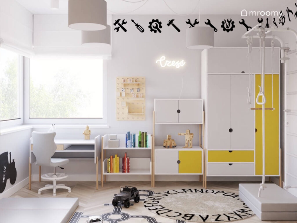 Jasnoszara ściana w pokoju chłopca a na niej szaro żółte meble na drewnianych nogach naklejki w kształcie narzędzi oraz ledon a na podłodze dywan z alfabetem