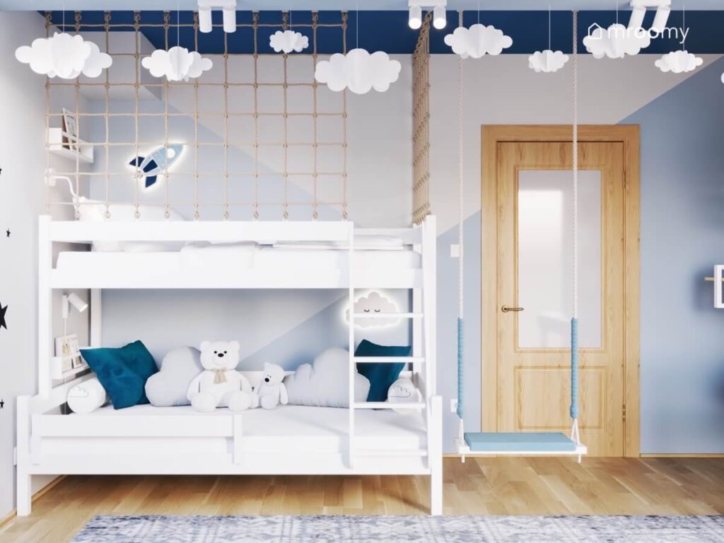 Białe łóżko zabezpieczone siatką i oświetlone uroczymi lampkami na środku huśtawka a u sufitu chmurki w pokoju dla chłopca