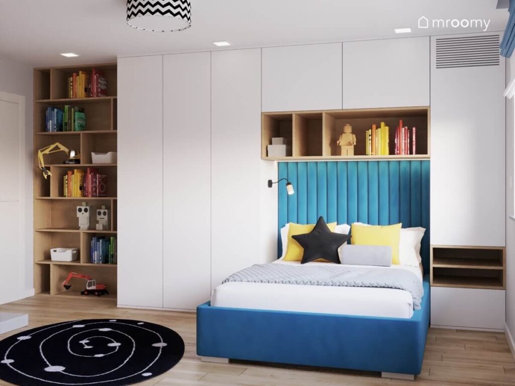 Strefa spania w pokoju chłopca z dużym niebieskim tapicerowanym łóżkiem uzupełnionym miękkimi panelami a dookoła białe szafy i drewniany regał