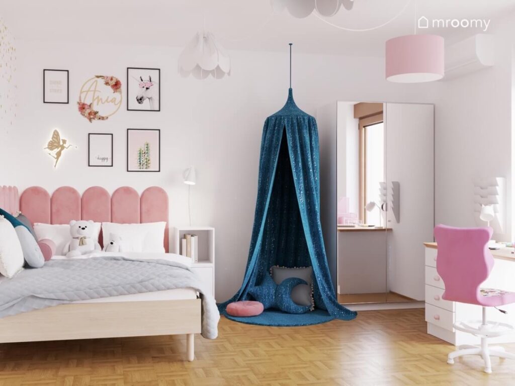 Drewniane łóżko uzupełnione różowymi panelami obok kącik z niebieskim baldachimem w pokoju dziewczynki a na ścianie obrazki w ramkach