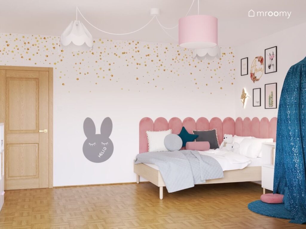 Biały pokój dla dziewczynki z drewnianym łóżkiem uzupełnionym różowymi panelami tapetą w złote gwiazdki oraz tablicą kredową w kształcie królika