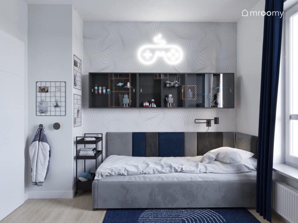 Strefa spania w pokoju chłopca z szarym tapicerowanym łóżkiem uzupełnionym miękkimi panelami ledonem w kształcie pada i minimalistyczną tapetą