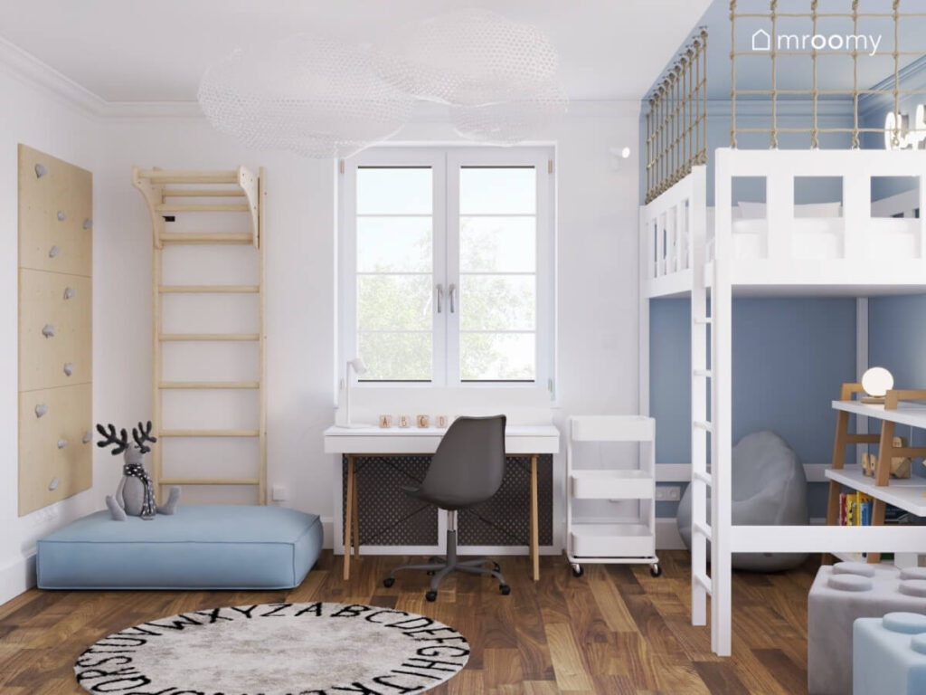 Biały pokój dla chłopca ze ścianką wspinaczkową drabinką gimnastyczną i niebieskimi pufami a także białym biurkiem na drewnianych nogach i niebieską strefą spania