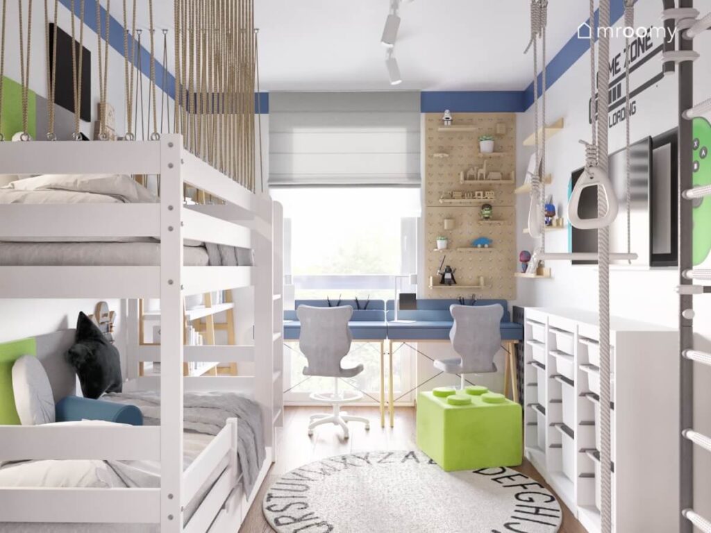 Jasny pokój dwóch chłopców z białym łóżkiem piętrowym białym regałem oraz niebieskimi biurkami i dywanem z alfabetem
