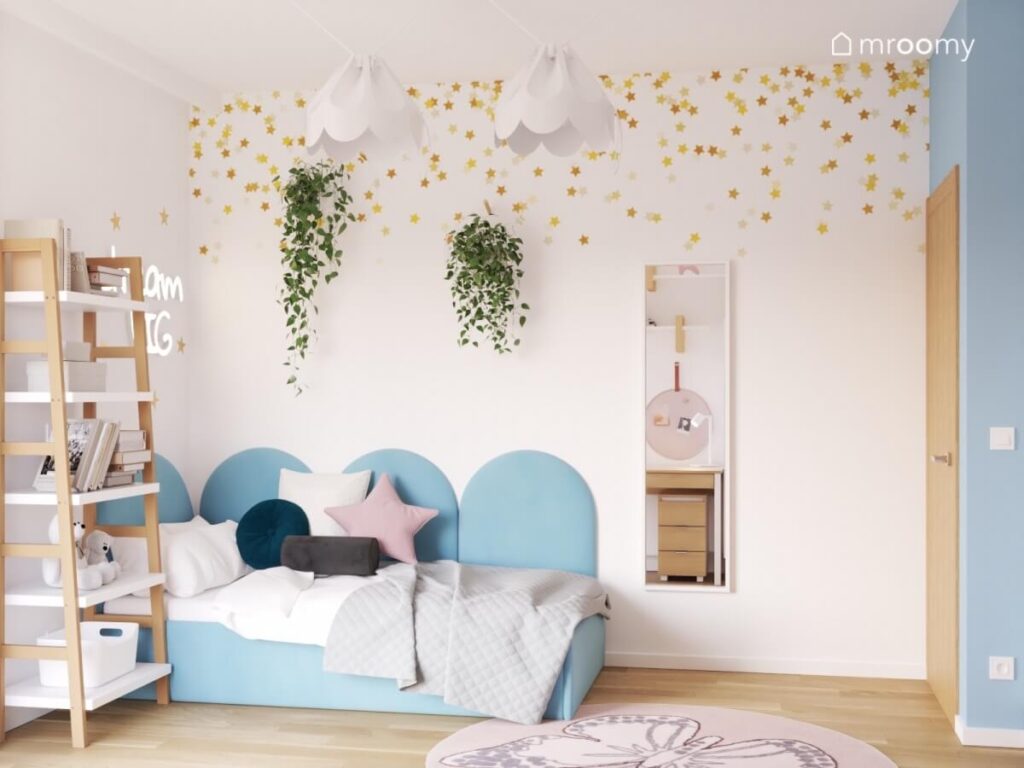 Strefa spania w pokoju dla dziewczynki z niebieskim tapicerowanym łóżkiem nad którym wisi ledon kwietniki oraz lustro na ścianie złote gwiazdki a na suficie lampy bezy