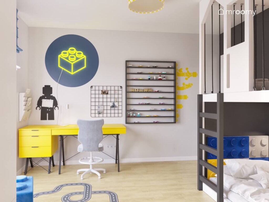 Strefa nauki w pokoju chłopca z żółtym biurkiem z kontenerkiem oraz tablicą kredową w kształcie ludzika Lego a obok półka na klocki i samochody