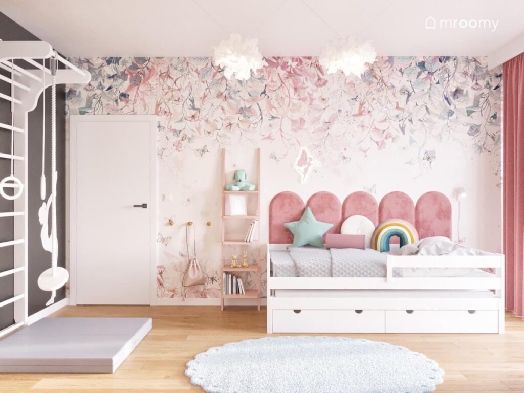 Białe łóżko w pokoju dziewczynki uzupełnione różowymi panelami a na ścianie tapeta w kwiaty