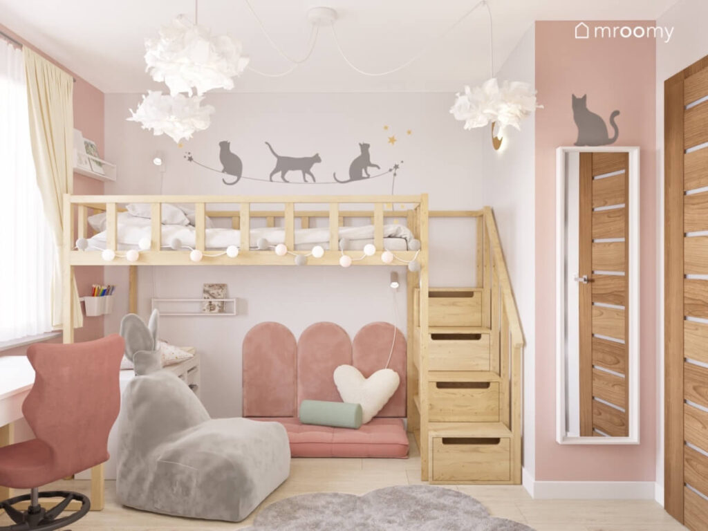 Drewniana antresola z miejscem do spania ozdobiona girlandą cotton balls na ścianach naklejki kotki a na podłodze szara pufa królik w pokoju dziewczynki