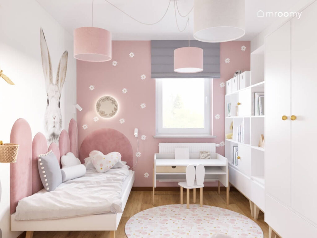 Białe łóżko uzupełnione miękkimi różowymi panelami nad nim naklejka królik obok biurko z szufladą a po przeciwnej stronie białe regały w pokoju dla dziewczynki