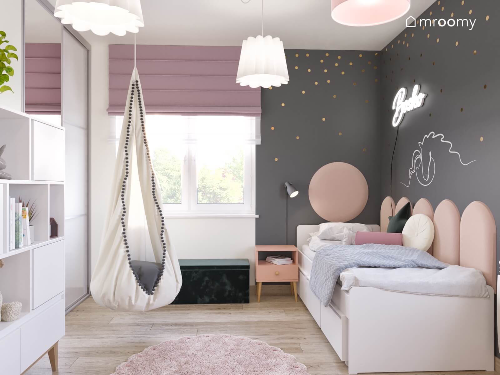 Szara strefa spania w pokoju dziewczynki a w niej łóżko uzupełnione panelami ściennymi a nad nim ledon naklejka z koniem i złote kropki obok wiszący fotel kokon